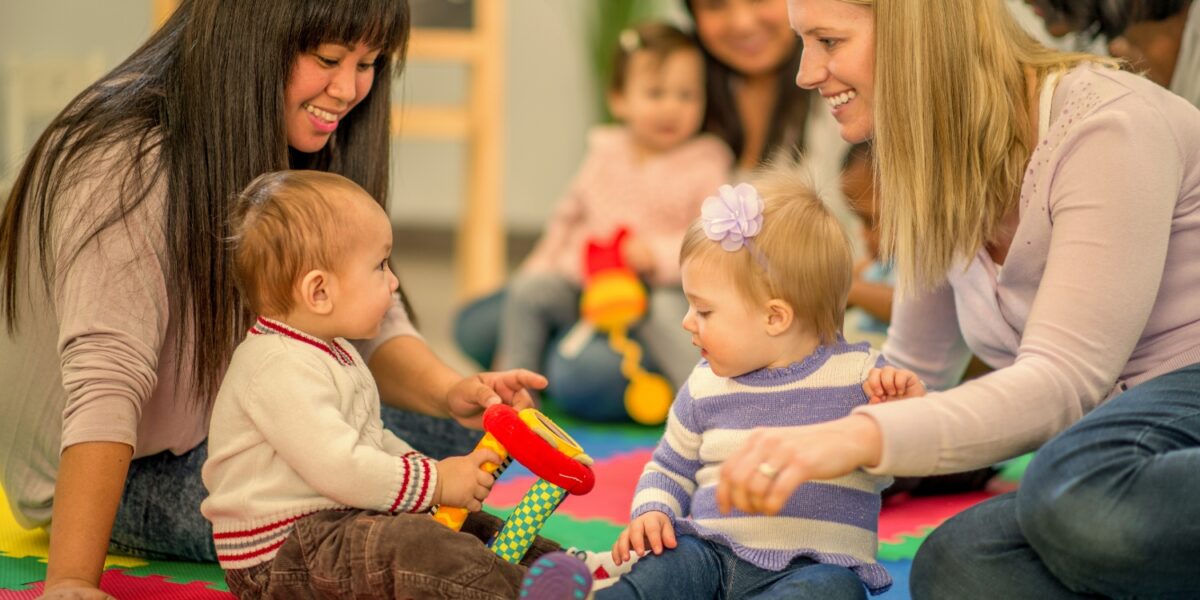 Zwei Frauen sitzen auf einer bunten Schaumstoffmatte mit zwei Babys, die mit Spielzeug spielen, während im Hintergrund andere Erwachsene und Kinder zu sehen sind.