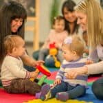 Zwei Frauen sitzen auf einer bunten Schaumstoffmatte mit zwei Babys, die mit Spielzeug spielen, während im Hintergrund andere Erwachsene und Kinder zu sehen sind.