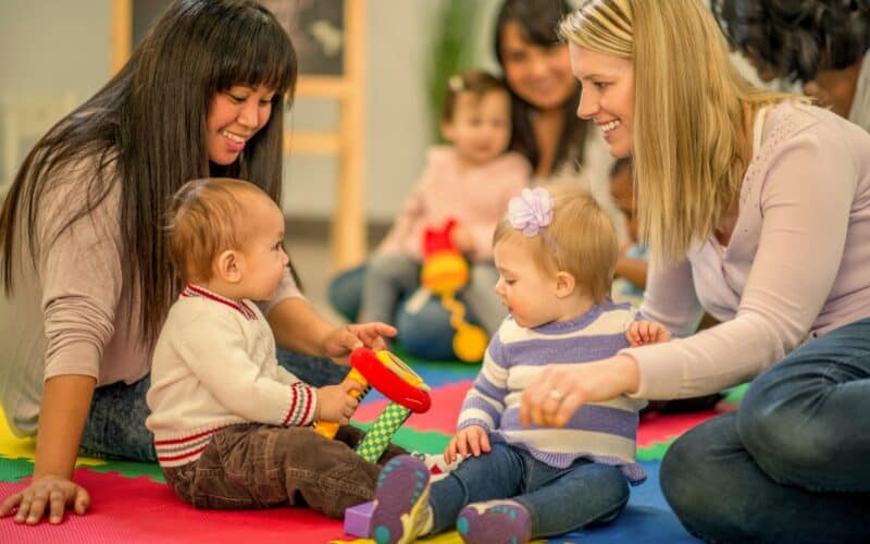 Zwei Frauen sitzen auf einer bunten Schaumstoffmatte mit zwei Babys, die mit Spielzeug spielen, während im Hintergrund andere Erwachsene und Kinder zu sehen sind.