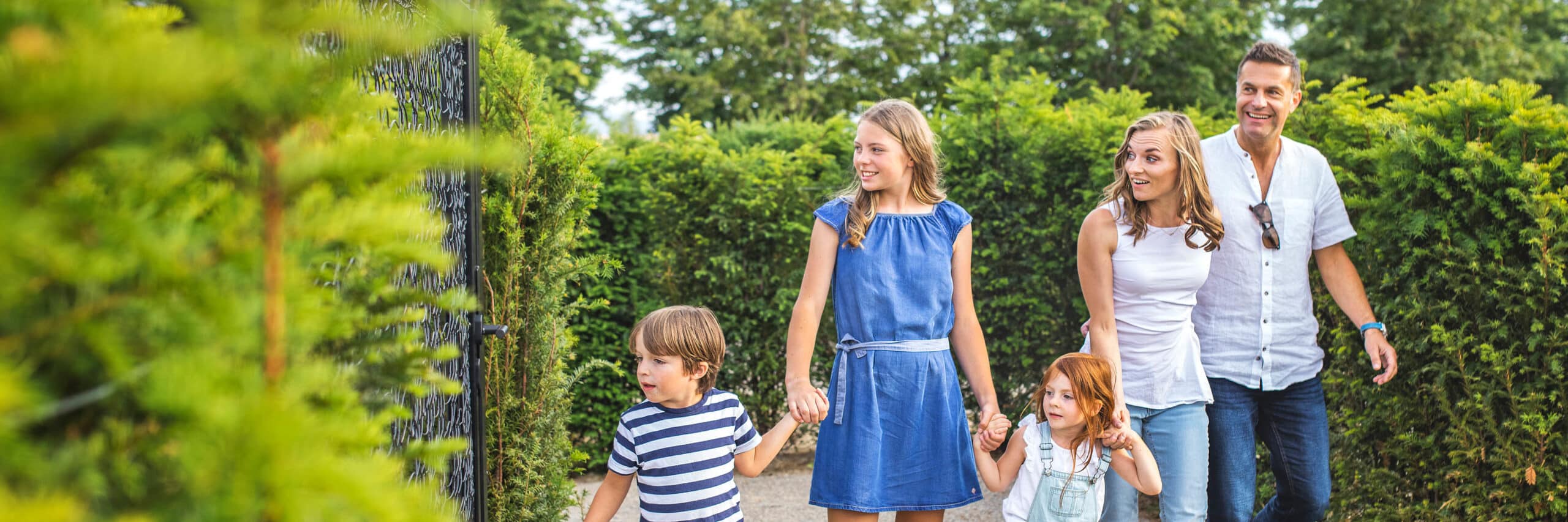 Eine fünfköpfige Familie, darunter zwei Erwachsene und drei Kinder, geht an einem sonnigen Tag gemeinsam auf einem von Hecken gesäumten Weg spazieren.