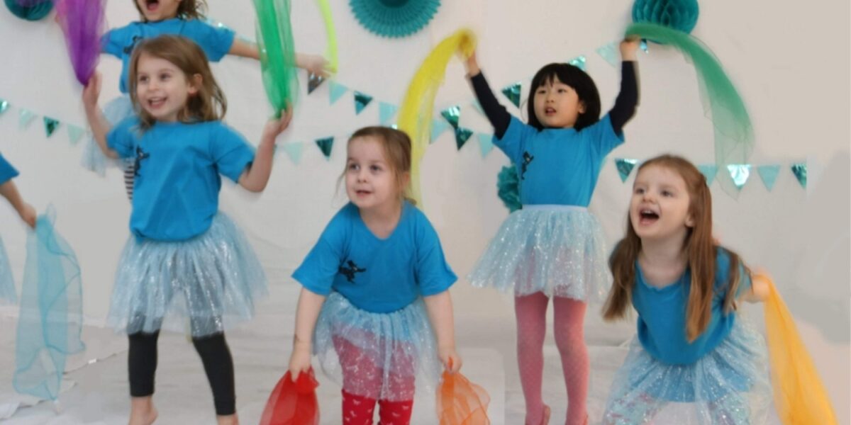 Fünf kleine Kinder in blauen Hemden und Röcken halten bunte Tücher und tanzen in einem geschmückten Raum mit türkisfarbenen Papierdekorationen und Wimpeln an der Wand.