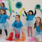 Fünf kleine Kinder in blauen Hemden und Röcken halten bunte Tücher und tanzen in einem geschmückten Raum mit türkisfarbenen Papierdekorationen und Wimpeln an der Wand.