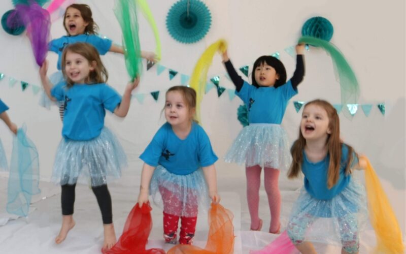 Fünf kleine Kinder in blauen Hemden und Röcken halten bunte Tücher und tanzen in einem geschmückten Raum mit türkisfarbenen Papierdekorationen und Wimpeln an der Wand.