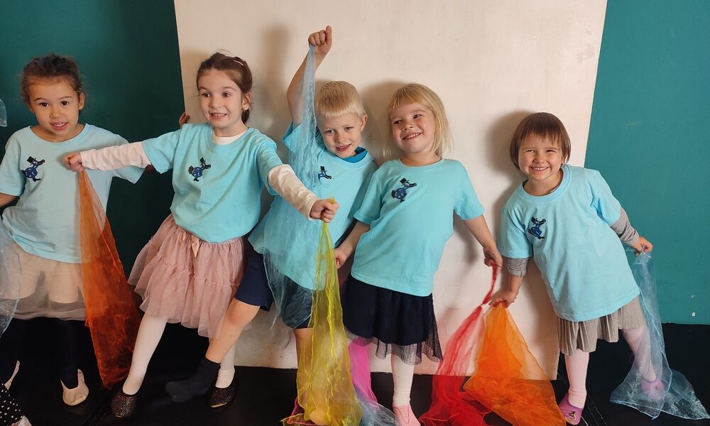 Fünf kleine Kinder in passenden blauen T-Shirts stehen in einer Reihe, halten bunte Schals in der Hand und lächeln vor einem türkisfarbenen und weißen Hintergrund.