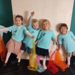 Fünf kleine Kinder in passenden blauen T-Shirts stehen in einer Reihe, halten bunte Schals in der Hand und lächeln vor einem türkisfarbenen und weißen Hintergrund.