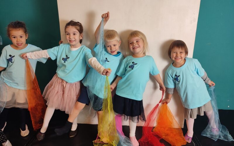 Fünf kleine Kinder in passenden blauen T-Shirts stehen in einer Reihe, halten bunte Schals in der Hand und lächeln vor einem türkisfarbenen und weißen Hintergrund.