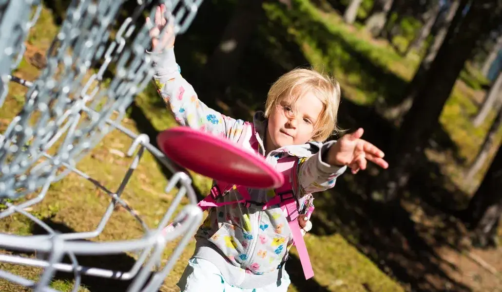 Ein kleines Kind in einer gemusterten Jacke wirft eine rosafarbene Scheibe auf einen Disc-Golf-Korb aus Metall in einem sonnenbeschienenen Waldgebiet.