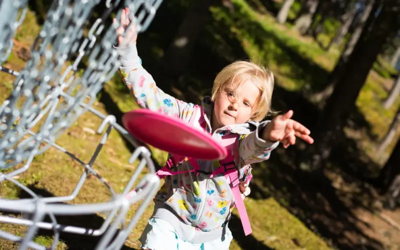 Ein kleines Kind in einer gemusterten Jacke wirft eine rosafarbene Scheibe auf einen Disc-Golf-Korb aus Metall in einem sonnenbeschienenen Waldgebiet.