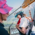 Eine Person, die einen Helm und ein Gurtzeug trägt, bereitet sich auf eine Seilrutschenfahrt vor, während eine andere Person mit einem rosa Hut in der Nähe steht. Im Hintergrund sind Berge und grüne Landschaften zu sehen.