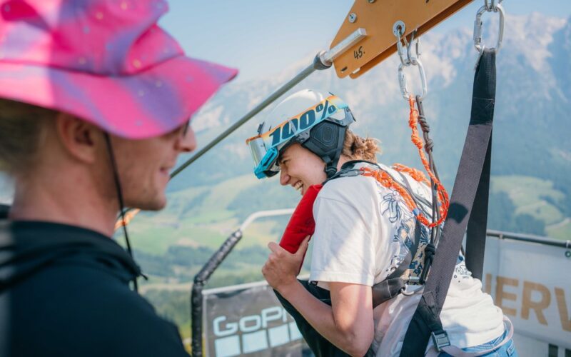Eine Person, die einen Helm und ein Gurtzeug trägt, bereitet sich auf eine Seilrutschenfahrt vor, während eine andere Person mit einem rosa Hut in der Nähe steht. Im Hintergrund sind Berge und grüne Landschaften zu sehen.