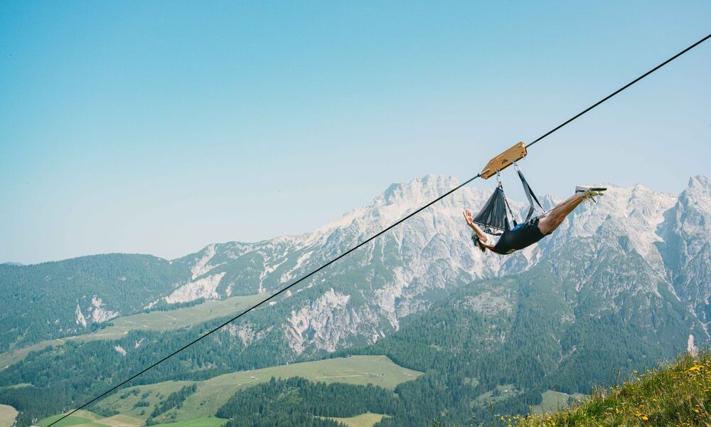 Eine Person fährt auf einer Seilrutsche hoch über einem grünen Tal mit Bergen im Hintergrund unter einem klaren blauen Himmel.
