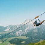 Eine Person fährt auf einer Seilrutsche hoch über einem grünen Tal mit Bergen im Hintergrund unter einem klaren blauen Himmel.