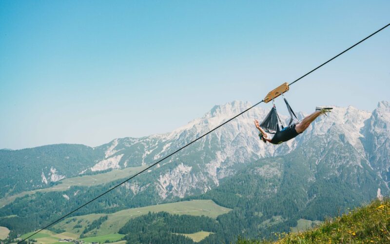 Eine Person fährt auf einer Seilrutsche hoch über einem grünen Tal mit Bergen im Hintergrund unter einem klaren blauen Himmel.