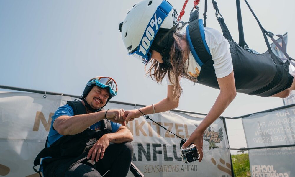 Eine Frau mit Helm und Gurtzeug ist horizontal aufgehängt, während sie einen lächelnden Mann in Outdoor-Ausrüstung mit der Faust anstößt; im Hintergrund ist ein Transparent zu sehen.
