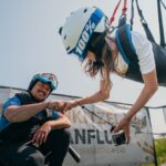 Eine Frau mit Helm und Gurtzeug ist horizontal aufgehängt, während sie einen lächelnden Mann in Outdoor-Ausrüstung mit der Faust anstößt; im Hintergrund ist ein Transparent zu sehen.