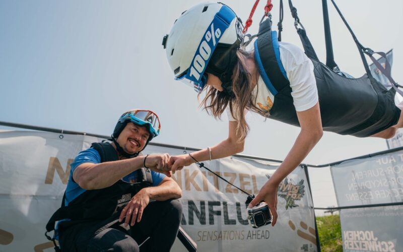 Eine Frau mit Helm und Gurtzeug ist horizontal aufgehängt, während sie einen lächelnden Mann in Outdoor-Ausrüstung mit der Faust anstößt; im Hintergrund ist ein Transparent zu sehen.
