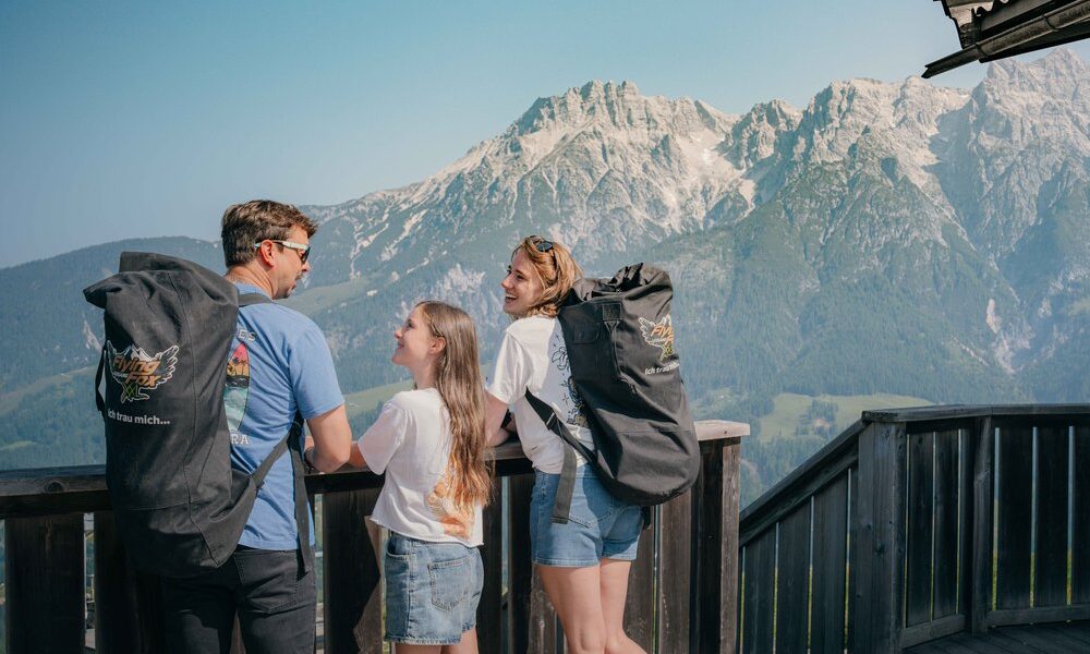 Drei Personen mit Rucksäcken stehen auf einem Holzbalkon mit Blick auf eine malerische Gebirgskette unter einem strahlend blauen Himmel.
