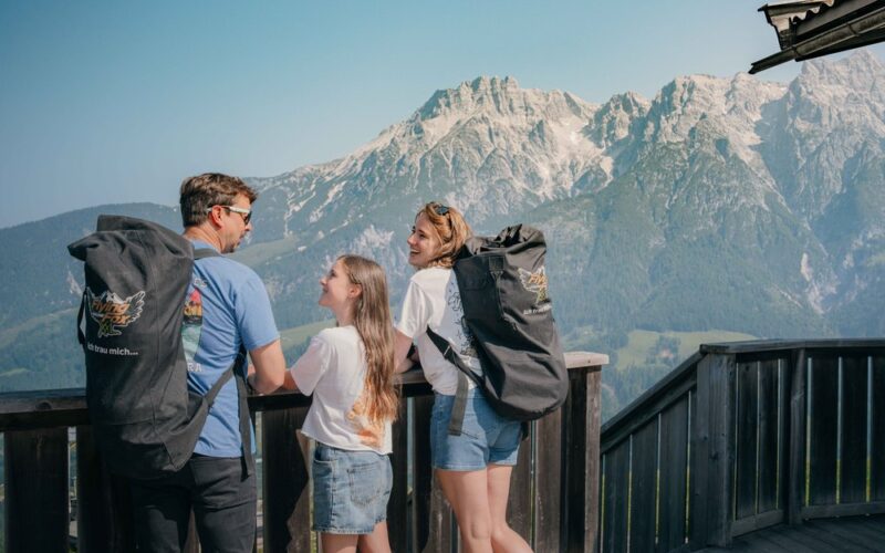 Drei Personen mit Rucksäcken stehen auf einem Holzbalkon mit Blick auf eine malerische Gebirgskette unter einem strahlend blauen Himmel.