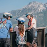 Drei Personen mit Helmen und Klettergurten stehen auf einem Holzdeck und blicken an einem klaren Tag auf eine Berglandschaft.