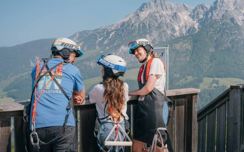 Drei Personen mit Helmen und Klettergurten stehen auf einem Holzdeck und blicken an einem klaren Tag auf eine Berglandschaft.