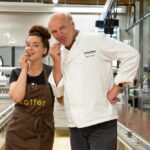 Zwei Personen, eine in einer braunen Schürze und die andere in einer weißen Kochjacke, stehen in einer Fabrikhalle und probieren Schokolade von einem großen Tisch.