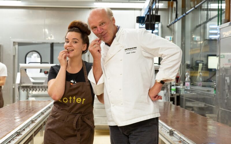 Zwei Personen, eine in einer braunen Schürze und die andere in einer weißen Kochjacke, stehen in einer Fabrikhalle und probieren Schokolade von einem großen Tisch.