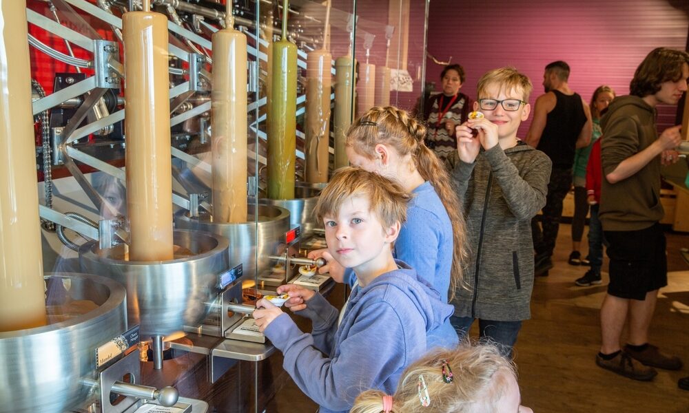 Kinder stehen an einer Station zur Herstellung von Süßigkeiten und beobachten Maschinen, die flüssige Süßigkeiten in Formen füllen. Erwachsene und andere Kinder sind im Hintergrund zu sehen.