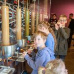 Kinder stehen an einer Station zur Herstellung von Süßigkeiten und beobachten Maschinen, die flüssige Süßigkeiten in Formen füllen. Erwachsene und andere Kinder sind im Hintergrund zu sehen.