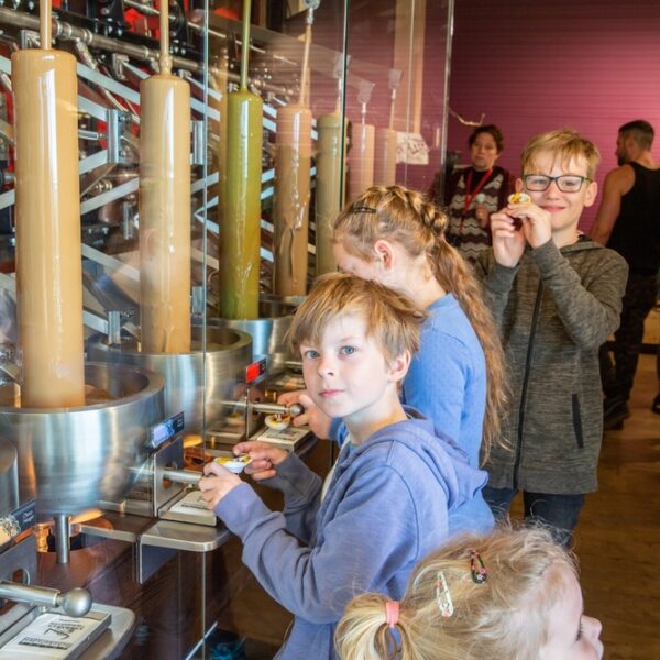 Kinder stehen an einer Station zur Herstellung von Süßigkeiten und beobachten Maschinen, die flüssige Süßigkeiten in Formen füllen. Erwachsene und andere Kinder sind im Hintergrund zu sehen.