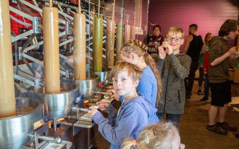 Kinder stehen an einer Station zur Herstellung von Süßigkeiten und beobachten Maschinen, die flüssige Süßigkeiten in Formen füllen. Erwachsene und andere Kinder sind im Hintergrund zu sehen.