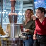 Drei Frauen stehen um einen Schokoladenbrunnen herum; eine Frau in einem roten Hemd probiert mit einem Löffel Schokolade und lächelt.