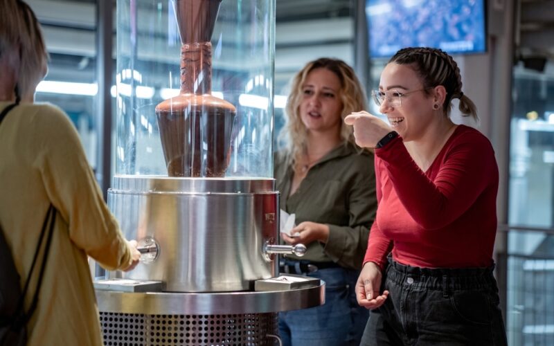 Drei Frauen stehen um einen Schokoladenbrunnen herum; eine Frau in einem roten Hemd probiert mit einem Löffel Schokolade und lächelt.