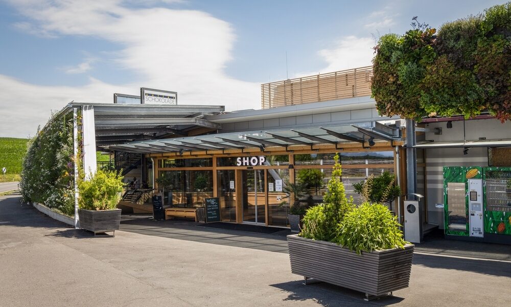 Modernes Schaufenster mit großen Glasfenstern mit der Aufschrift SHOP", Sitzgelegenheiten im Freien, Topfpflanzen und eine grüne vertikale Gartenwand unter einem teilweise bewölkten Himmel.