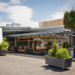 Modernes Schaufenster mit großen Glasfenstern mit der Aufschrift SHOP", Sitzgelegenheiten im Freien, Topfpflanzen und eine grüne vertikale Gartenwand unter einem teilweise bewölkten Himmel.