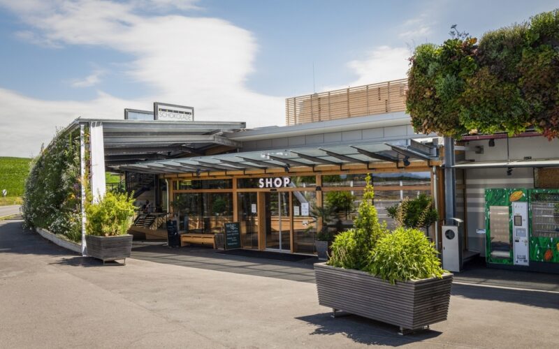 Modernes Schaufenster mit großen Glasfenstern mit der Aufschrift SHOP", Sitzgelegenheiten im Freien, Topfpflanzen und eine grüne vertikale Gartenwand unter einem teilweise bewölkten Himmel.