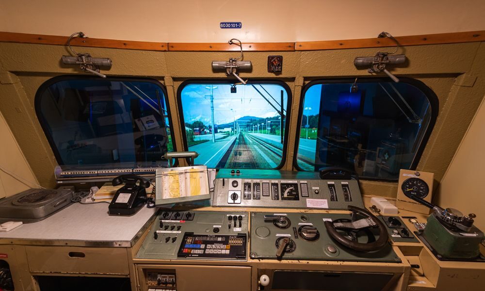 Blick aus dem Führerstand eines Zuges mit Schalttafeln, Hebeln, Messgeräten und einem Fenster mit Blick auf die Gleise und den Bahnsteig eines Bahnhofs.
