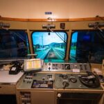 Blick aus dem Führerstand eines Zuges mit Schalttafeln, Hebeln, Messgeräten und einem Fenster mit Blick auf die Gleise und den Bahnsteig eines Bahnhofs.