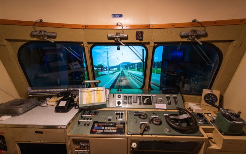 Blick aus dem Führerstand eines Zuges mit Schalttafeln, Hebeln, Messgeräten und einem Fenster mit Blick auf die Gleise und den Bahnsteig eines Bahnhofs.