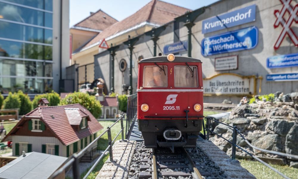 Eine rote Modelleisenbahn mit der Aufschrift "2095.11" fährt auf Außengleisen mit Miniaturgebäuden und Schildern im Hintergrund.