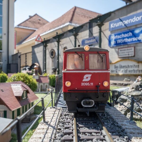 Eine rote Modelleisenbahn mit der Aufschrift "2095.11" fährt auf Außengleisen mit Miniaturgebäuden und Schildern im Hintergrund.