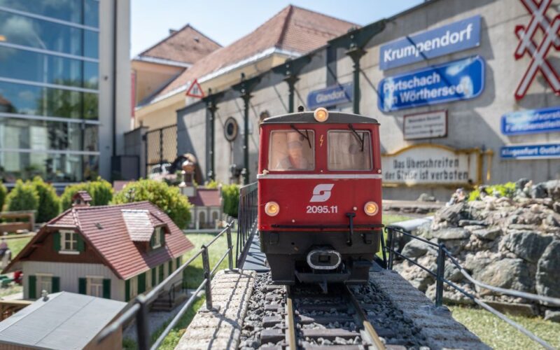 Eine rote Modelleisenbahn mit der Aufschrift "2095.11" fährt auf Außengleisen mit Miniaturgebäuden und Schildern im Hintergrund.