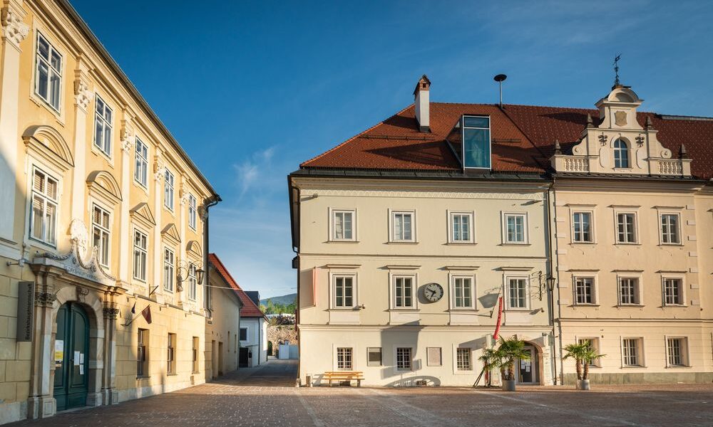 Ein ruhiger Stadtplatz mit zwei historischen Gebäuden, einer Bank, Palmen und einer Uhr an einer Fassade unter strahlend blauem Himmel.