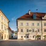 Ein ruhiger Stadtplatz mit zwei historischen Gebäuden, einer Bank, Palmen und einer Uhr an einer Fassade unter strahlend blauem Himmel.