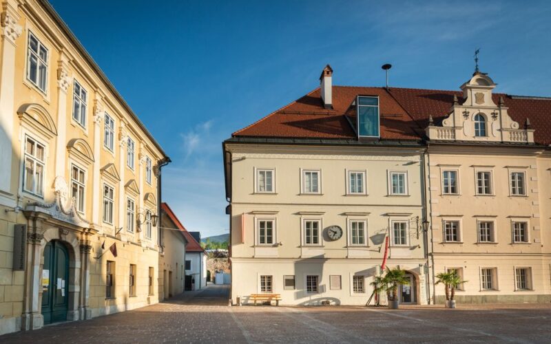 Ein ruhiger Stadtplatz mit zwei historischen Gebäuden, einer Bank, Palmen und einer Uhr an einer Fassade unter strahlend blauem Himmel.