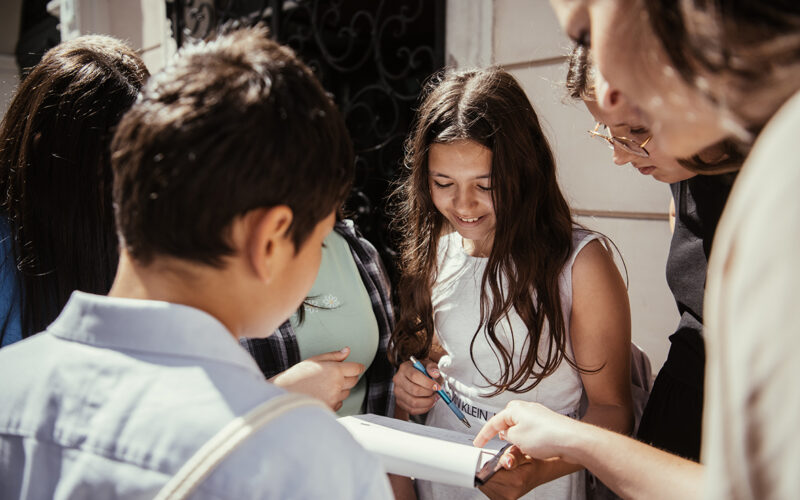 Eine Gruppe junger Menschen versammelt sich im Freien und schaut sich gemeinsam ein Klemmbrett oder ein Stück Papier an und schreibt darauf.
