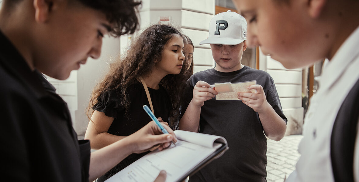 Vier Kinder stehen im Freien; eines schreibt auf einem Klemmbrett, ein anderes hält Papier, während zwei andere ihre Arbeit beobachten und gemeinsam diskutieren.