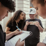 Vier Kinder stehen im Freien; eines schreibt auf einem Klemmbrett, ein anderes hält Papier, während zwei andere ihre Arbeit beobachten und gemeinsam diskutieren.