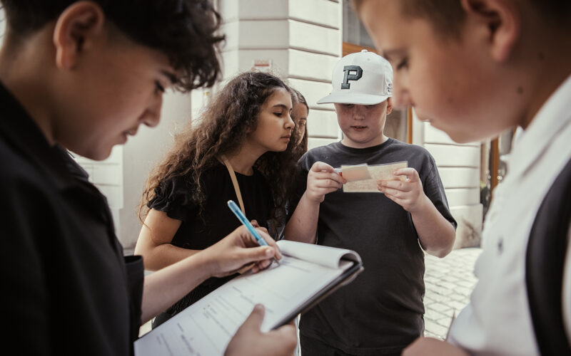 Vier Kinder stehen im Freien; eines schreibt auf einem Klemmbrett, ein anderes hält Papier, während zwei andere ihre Arbeit beobachten und gemeinsam diskutieren.