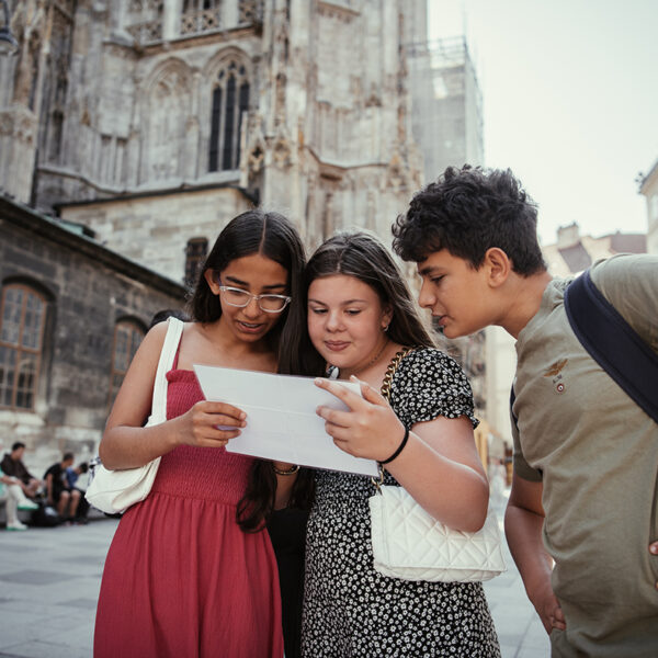 Drei Teenager stehen gemeinsam vor einem historischen Gebäude und schauen auf einen Tablet-Bildschirm.