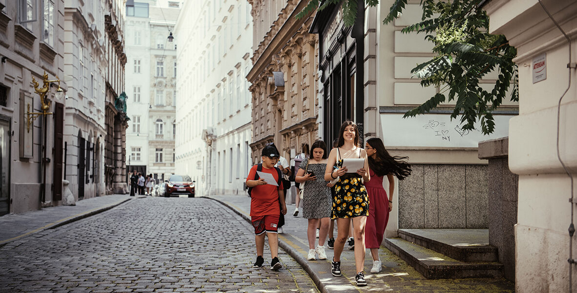 Vier junge Leute gehen an einem sonnigen Tag eine von historischen Gebäuden gesäumte Kopfsteinpflasterstraße entlang. Die Gruppe wirkt entspannt und in ein Gespräch vertieft.